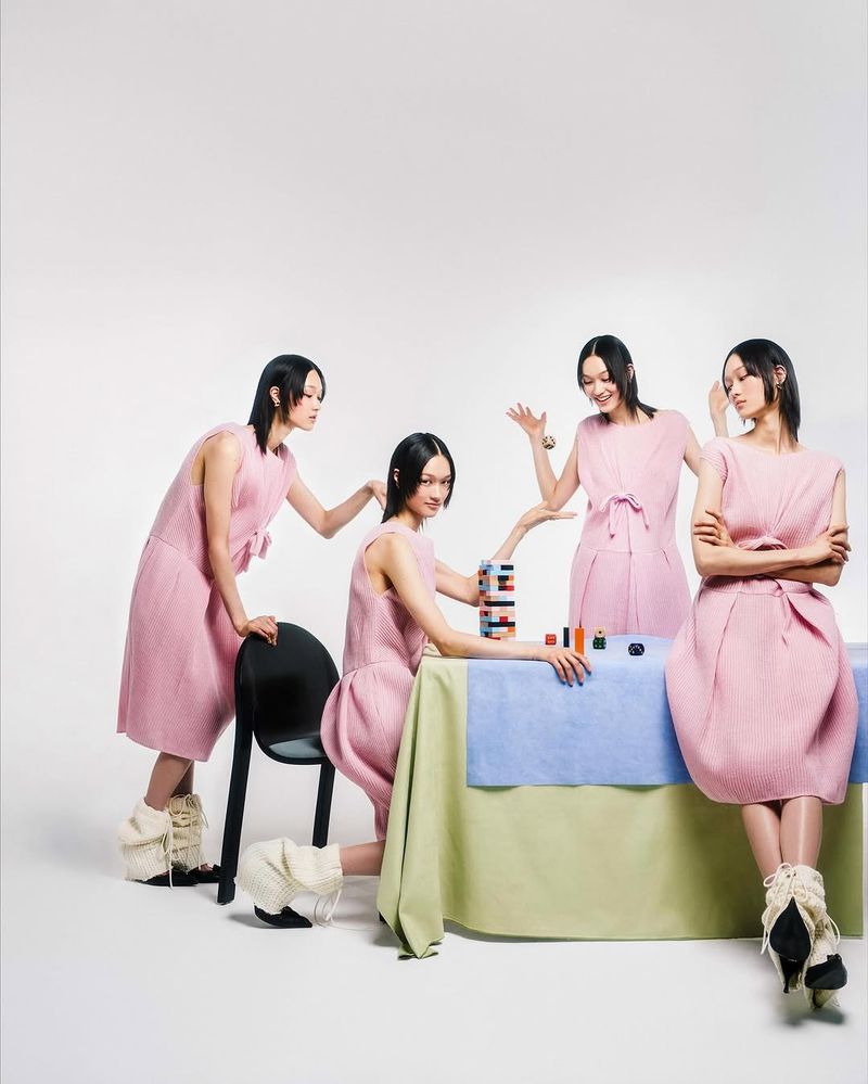 Four identical Asian women wearing pink dresses in a minimalist studio setting, seated around a table with colorful makeup and stacking toys.