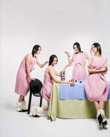 Four identical Asian women wearing pink dresses in a minimalist studio setting, seated around a table with colorful makeup and stacking toys.