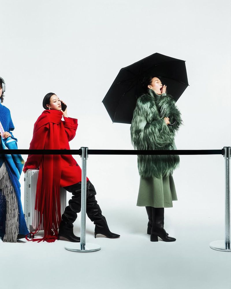 Three women in fashionable outerwear stand behind a black rope barrier in a studio setting, with one holding an umbrella and talking.