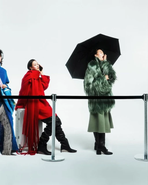 Three women in fashionable outerwear stand behind a black rope barrier in a studio setting, with one holding an umbrella and talking.