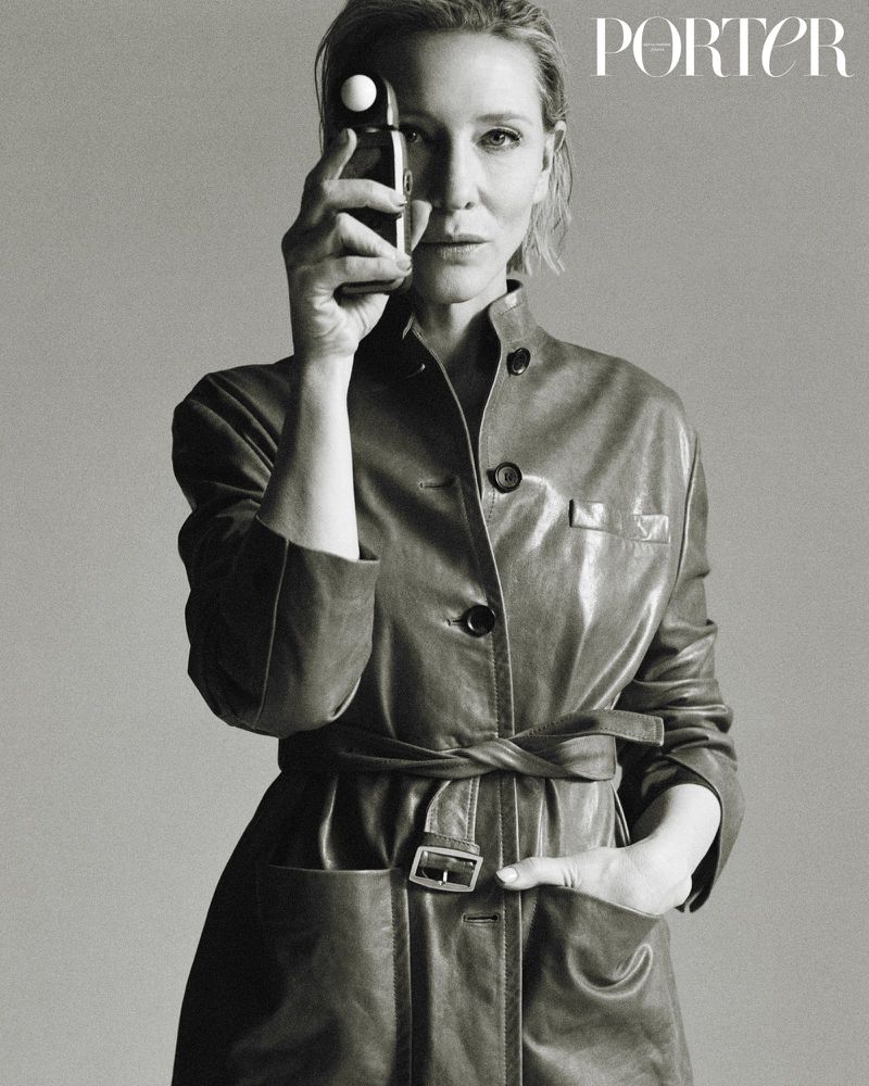 Black and white portrait of an adult woman holding a light meter to her eye while wearing a belted leather jacket in a studio.