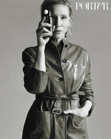 Black and white portrait of an adult woman holding a light meter to her eye while wearing a belted leather jacket in a studio.