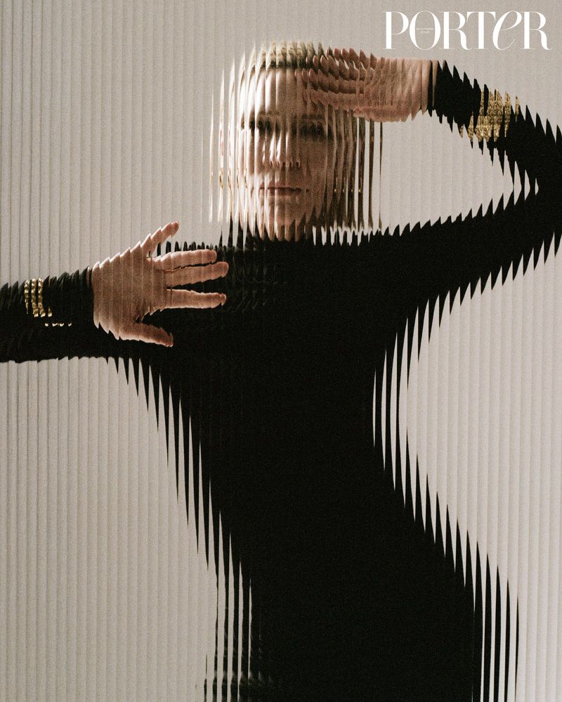 Fashion editorial shot of an adult woman in a black long-sleeve dress posing behind textured glass with the magazine logo in the upper corner.