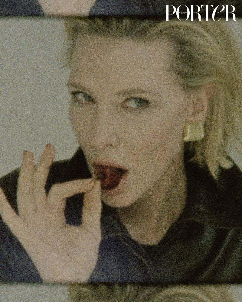 Close-up of a blonde woman holding a dark red fruit to her mouth, wearing a black collared shirt and gold earrings, shot indoors.