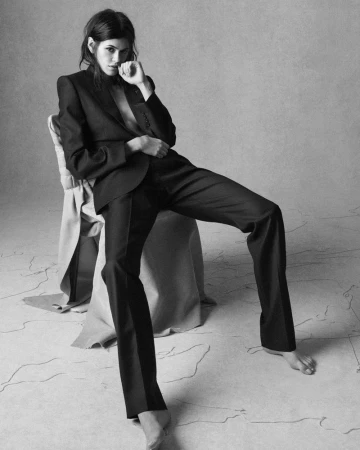 A barefoot woman in a dark suit sits in a photo studio, hand to face, with a neutral expression in black and white.