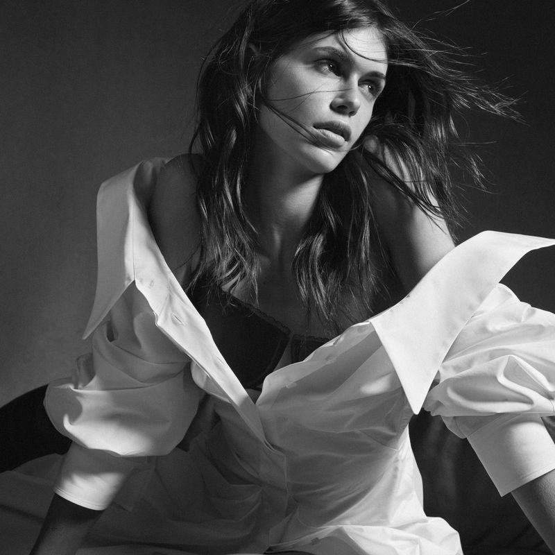 Monochrome studio shot of a young adult woman in a white button-down shirt, looking off-camera with wind-blown hair against a dark backdrop.