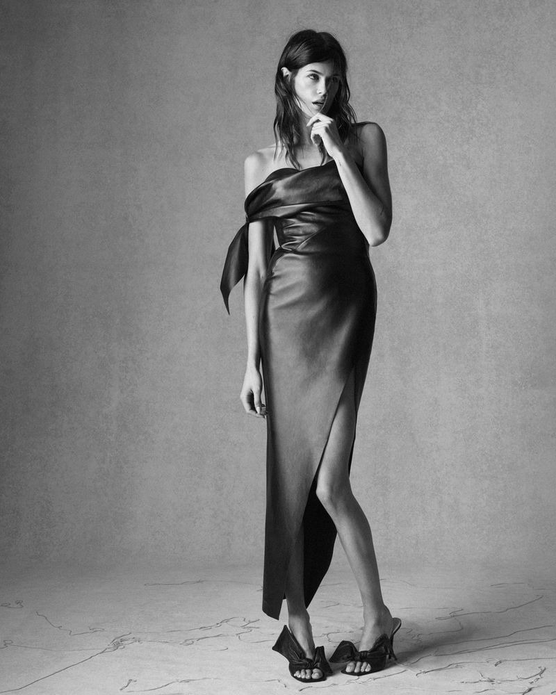 Monochrome studio shot of a woman posing in an off-the-shoulder midi dress with a thigh-high slit and high-heeled sandals with bow details.