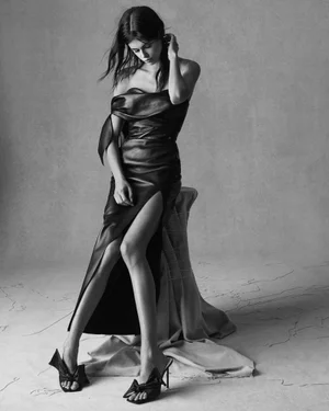 Model Kaia Gerber wearing a draped black leather gown with a high slit and bow-detail heeled mules in a studio portrait.