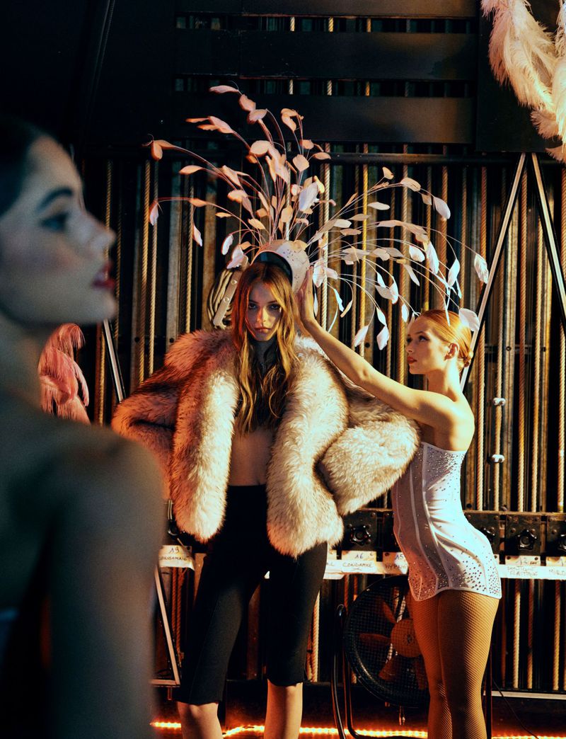 Three female models pose indoors wearing a fur coat, a feathered headdress, and a sequined dress in a dimly lit setting.