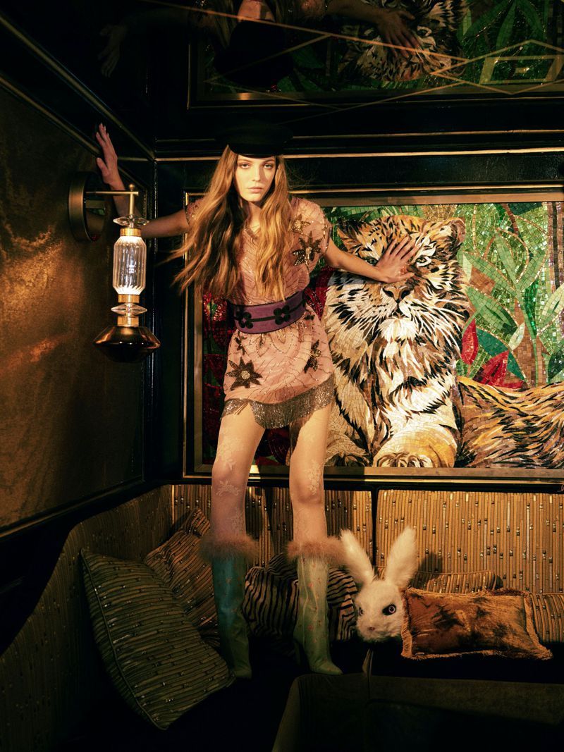 Fashion shot of a woman in a dress and hat posing next to a mosaic tiger artwork and a rabbit doll inside an ornate room.