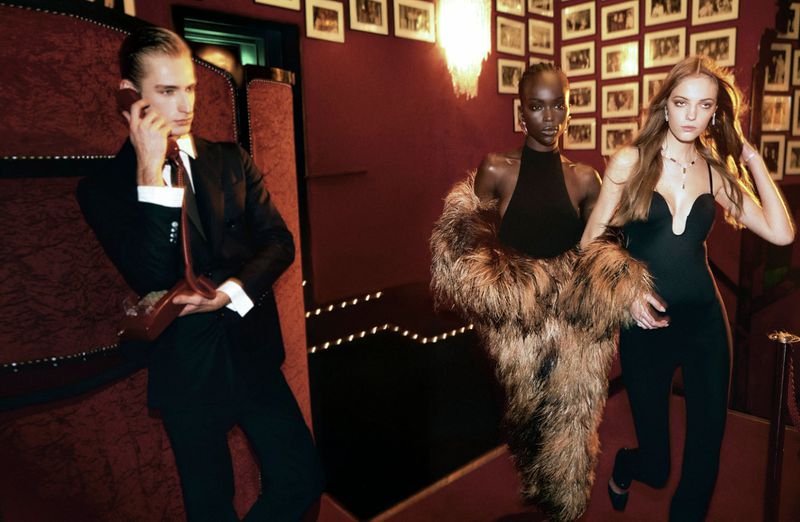 Elegant shot of two women and a man at an event. One woman wears a fur stole and the man talks on a phone in a dark, rich setting.
