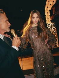 Woman in a sheer sequin dress standing next to a man holding a microphone in a dimly lit venue