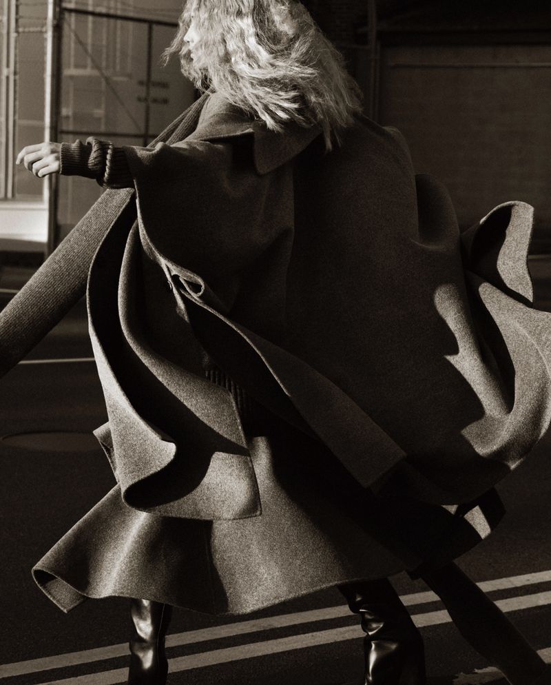 Woman in motion wearing oversized coat and black boots on a street in an editorial fashion photograph, capturing movement and style.