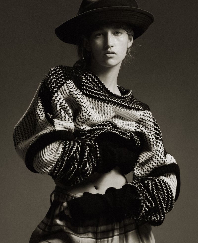 Monochrome portrait of a woman in a black hat and patterned sweater, striking a pose against a neutral backdrop, capturing a fashion editorial style.