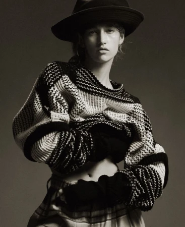 Monochrome portrait of a woman in a black hat and patterned sweater, striking a pose against a neutral backdrop, capturing a fashion editorial style.