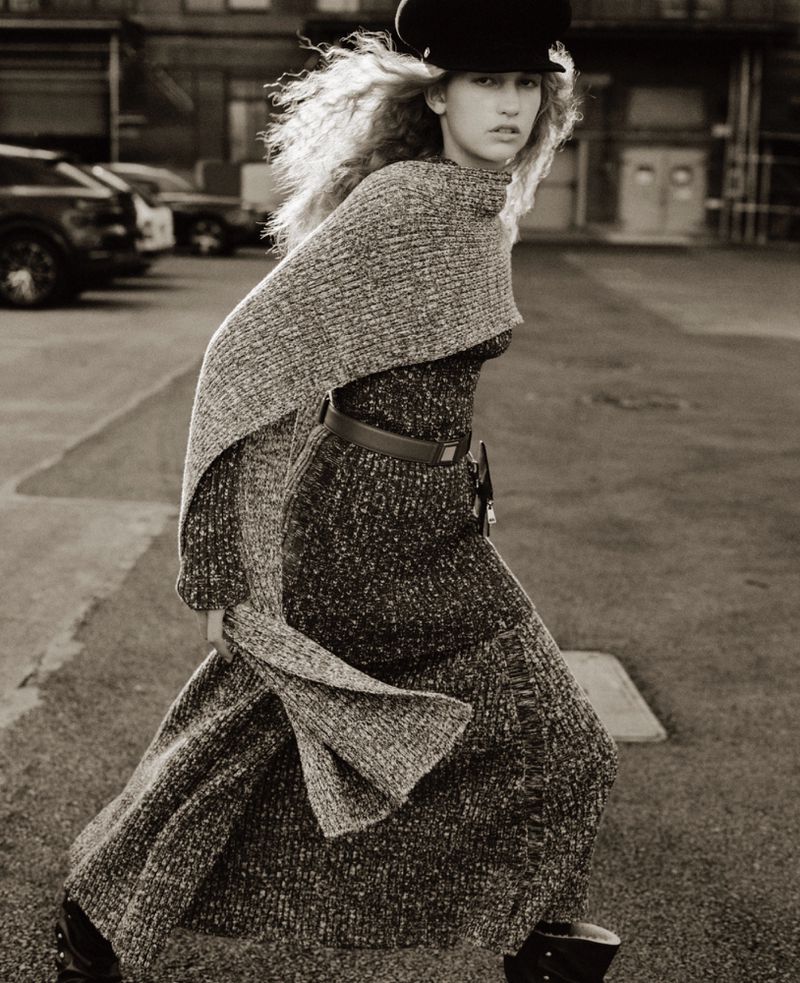 Model Apolline Rocco Fohrer walks outdoors wearing a grey textured knit dress, matching draped shawl, and black peaked cap.