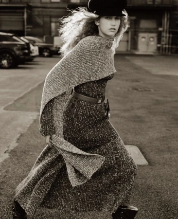 Model Apolline Rocco Fohrer walks outdoors wearing a grey textured knit dress, matching draped shawl, and black peaked cap.