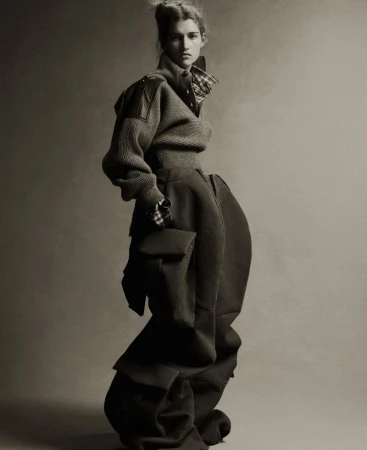 A woman in a fashion garment with a structured wool sweater and baggy pants poses in a studio against a plain backdrop.