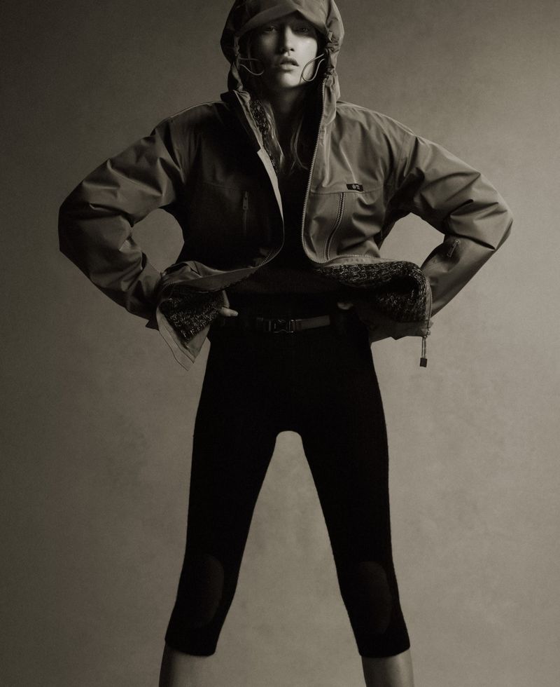 Model Apolline Rocco Fohrer posing with hands on hips wearing a hooded technical jacket and black leggings against a neutral backdrop.