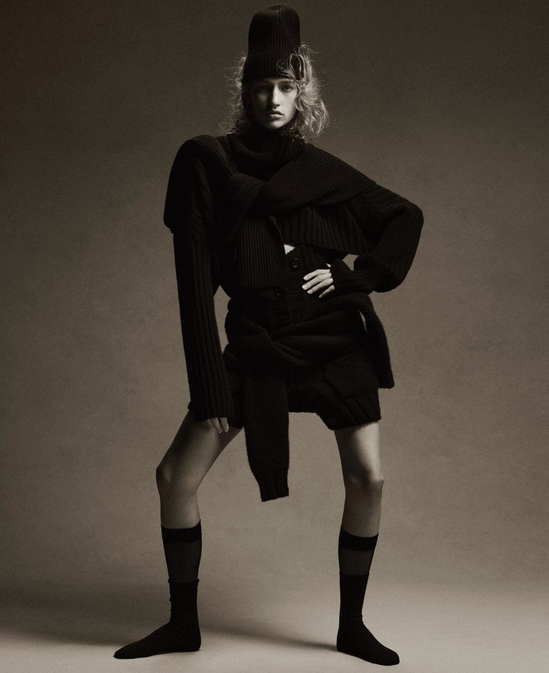 A striking full shot of a stylish adult female fashion model in an all-black knit outfit, standing against a neutral gradient backdrop in a studio setting.
