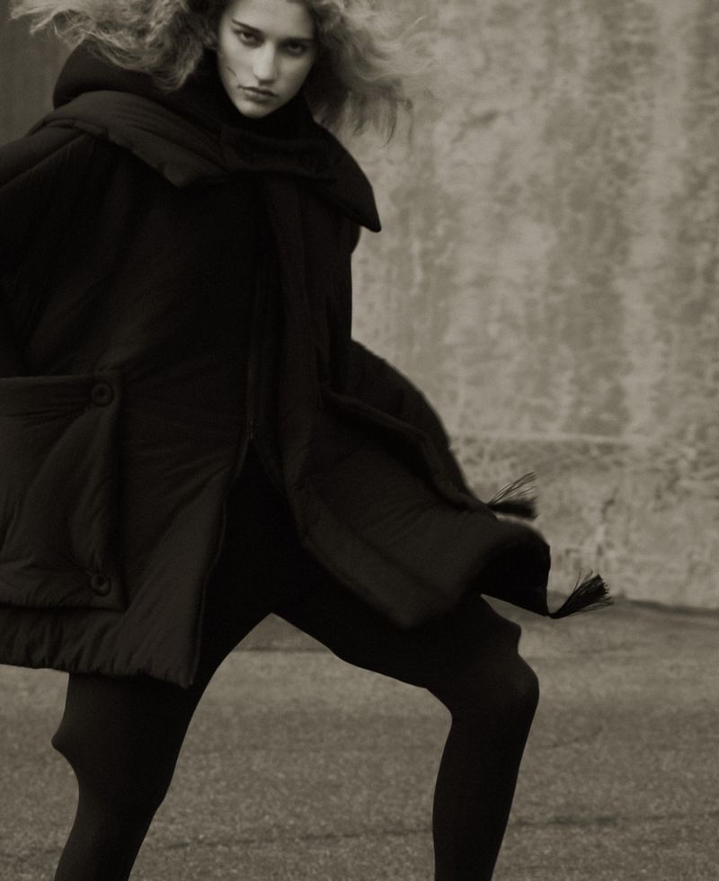 Fashionable woman wearing a dark hooded cape and leggings stands on a paved surface against a textured wall in a studio-style shot.