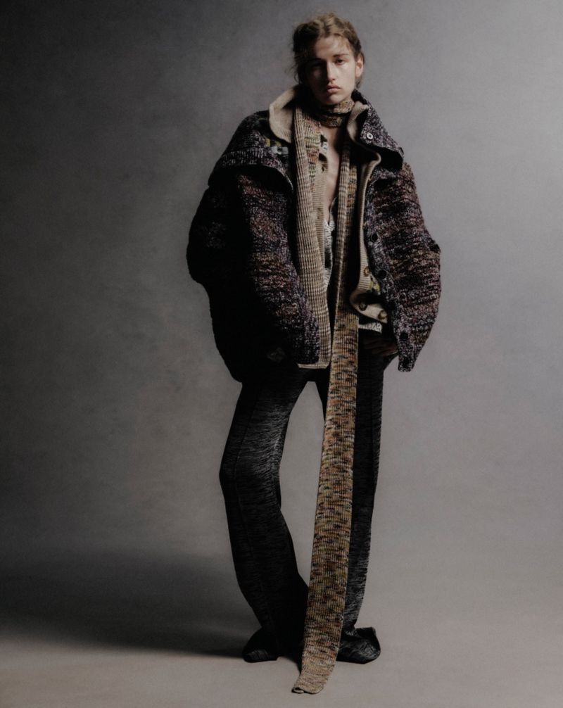 Fashionable adult stands in photo studio showcasing brown and black knit jacket, scarf, top, and pants against a gray backdrop.