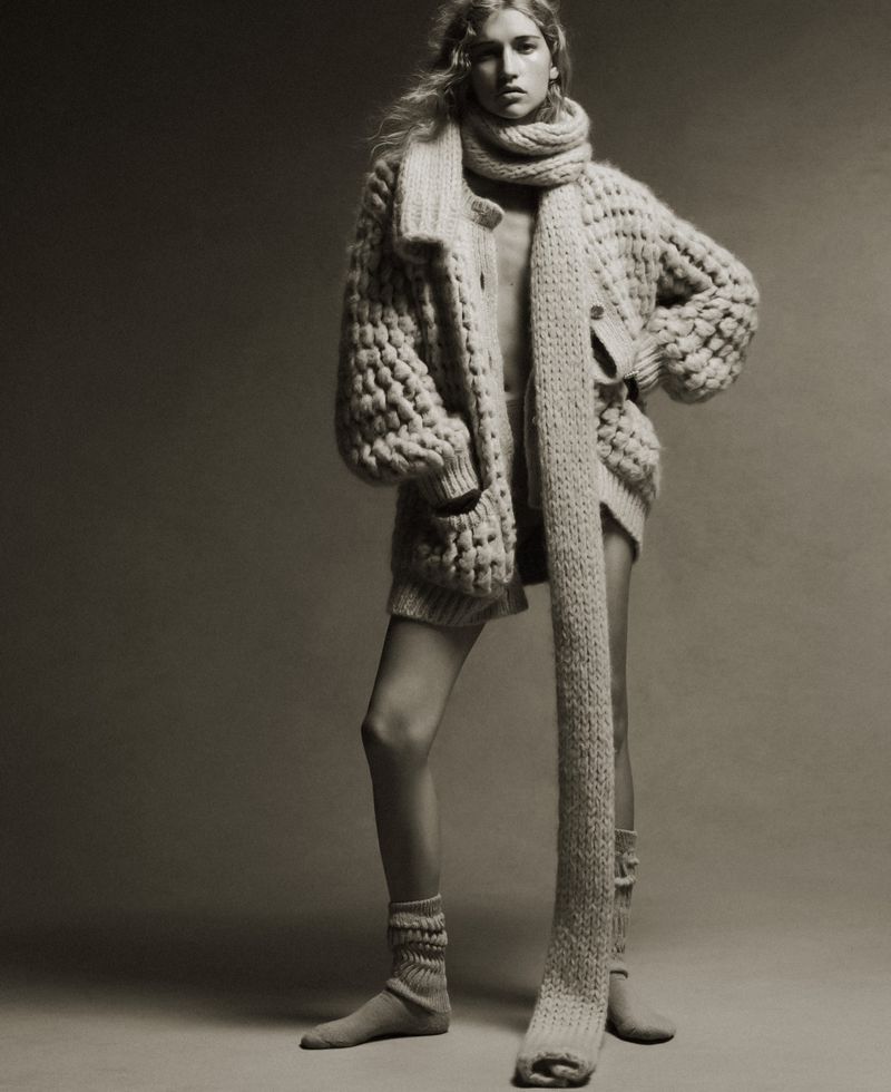 A blonde-haired woman in a bulky knit sweater and scarf poses indoors, her hands on her hips, wearing socks, against a grey backdrop.