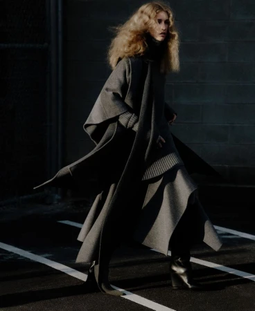A woman with blonde, curly hair walking outside, wearing a grey wool coat and black boots on pavement with white stripes.