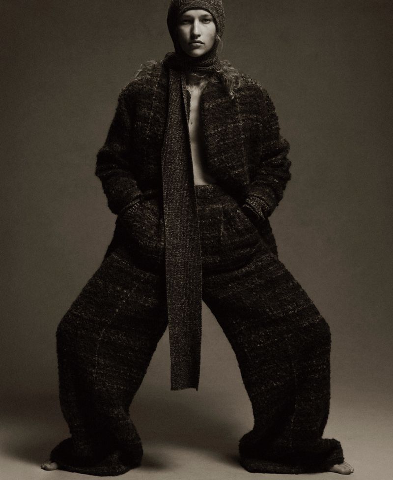 Fashionable adult female with knit hat and scarf wearing tweed coat and wide-leg tweed pants in a photo studio setting.