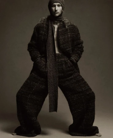 Fashionable adult female with knit hat and scarf wearing tweed coat and wide-leg tweed pants in a photo studio setting.