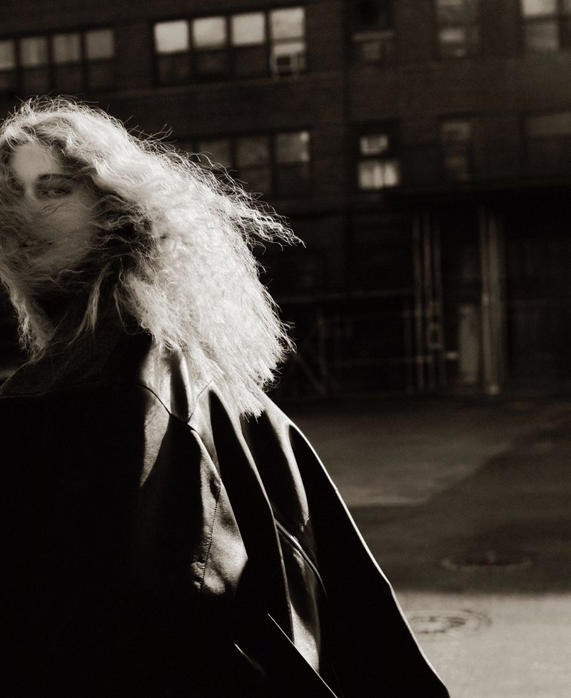 Black and white portrait of a woman with curly hair, wearing a jacket, standing in front of a building at night with visible windows.