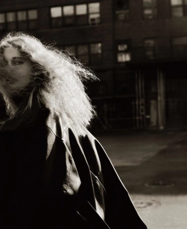 Black and white portrait of a woman with curly hair, wearing a jacket, standing in front of a building at night with visible windows.