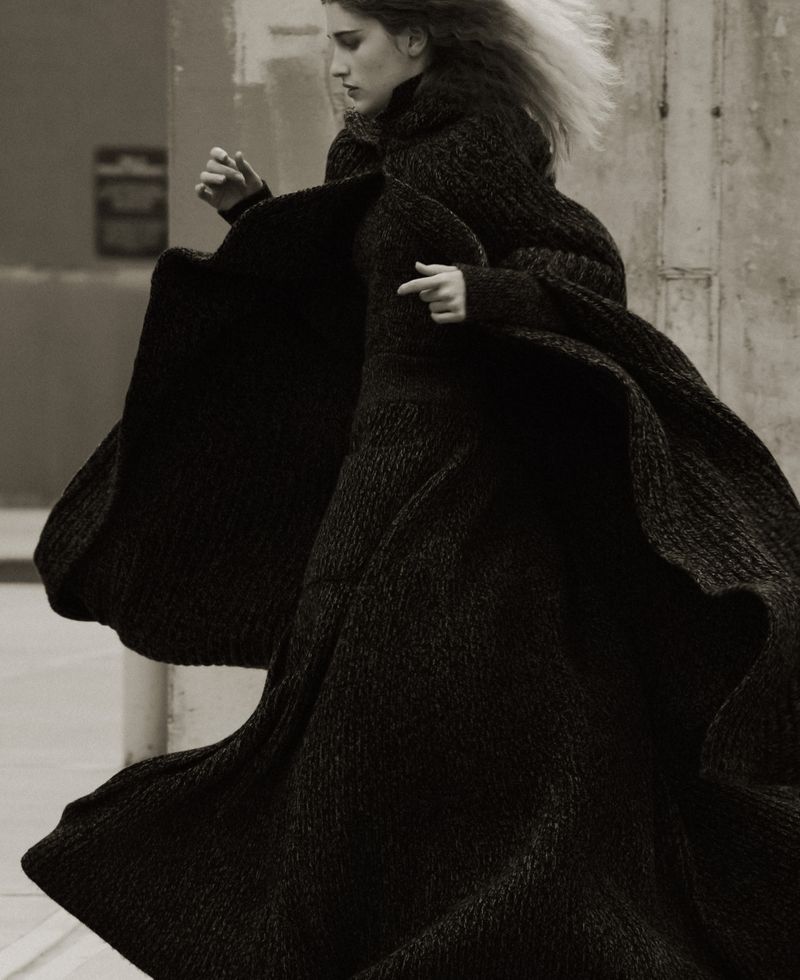 Fashion shot of a slender woman in a long, draped black coat, windswept hair, and contemplative expression against a neutral concrete backdrop.