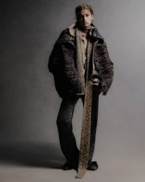 Model wearing layered knitwear and a long scarf against a neutral grey studio background