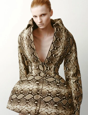 Fashionable adult female poses in a snakeskin-patterned dress in a photo studio, creating a striking, fashion-forward image with neutral tones.