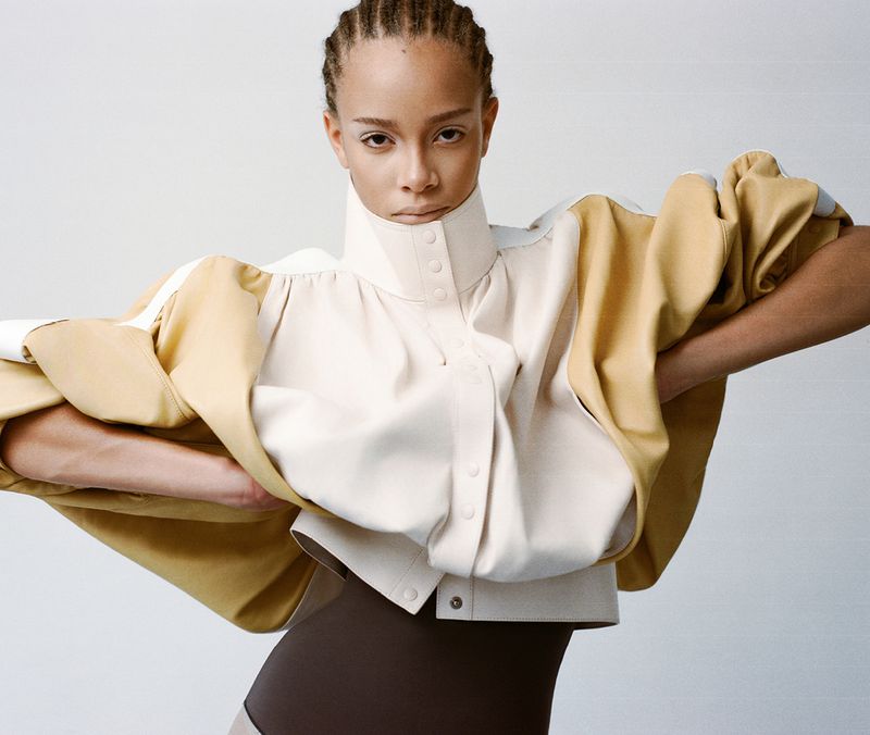 A light-skinned female poses indoors wearing a tan and cream jacket and brown bodysuit against a white background.