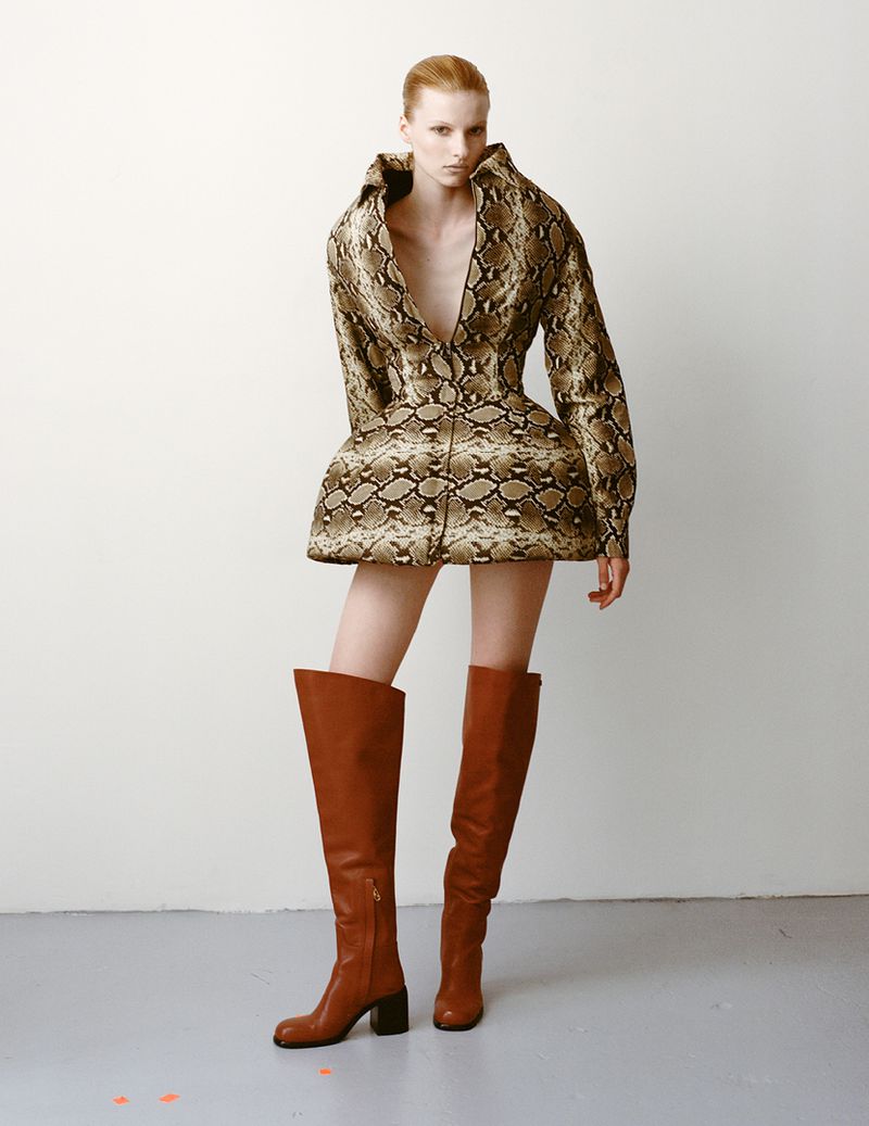 Fashion model in snakeskin-print mini dress and over-the-knee brown leather boots poses in a studio against a plain white wall.