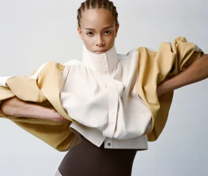 Model posing with hands on hips wearing a structured high-neck beige and cream cape jacket against a light studio backdrop.