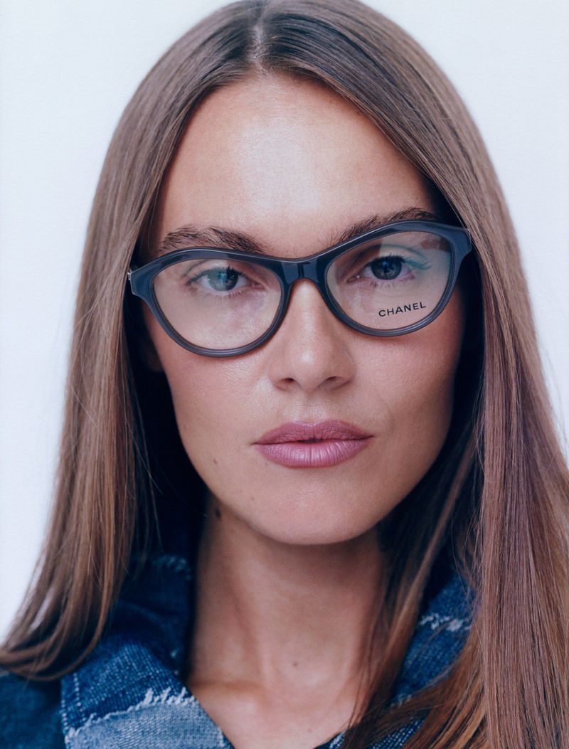 Close-up portrait of a woman with long hair wearing dark-framed eyeglasses with the Chanel logo, against a white backdrop.