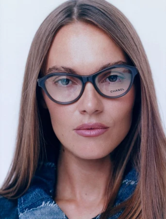 Close-up portrait of a woman with long hair wearing dark-framed eyeglasses with the Chanel logo, against a white backdrop.