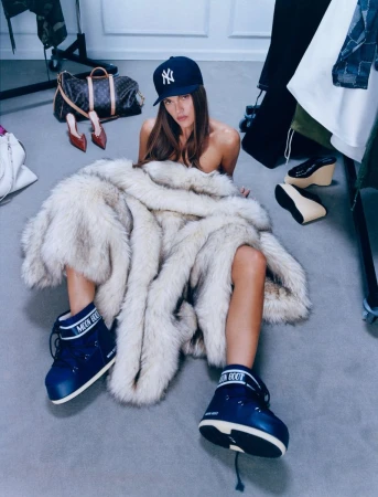 A woman in a fur coat and Moon Boots sits on the floor, wearing a baseball cap, with shoes and a bag in the background.