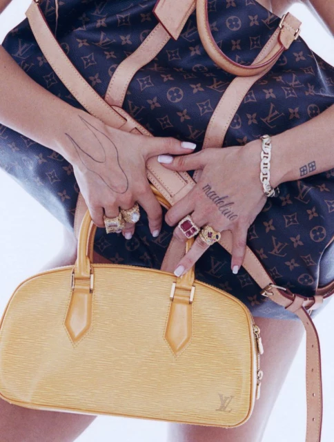 Close up of a woman's hands adorned with rings and tattoos, holding a yellow handbag with a brown monogrammed duffel bag in the background.