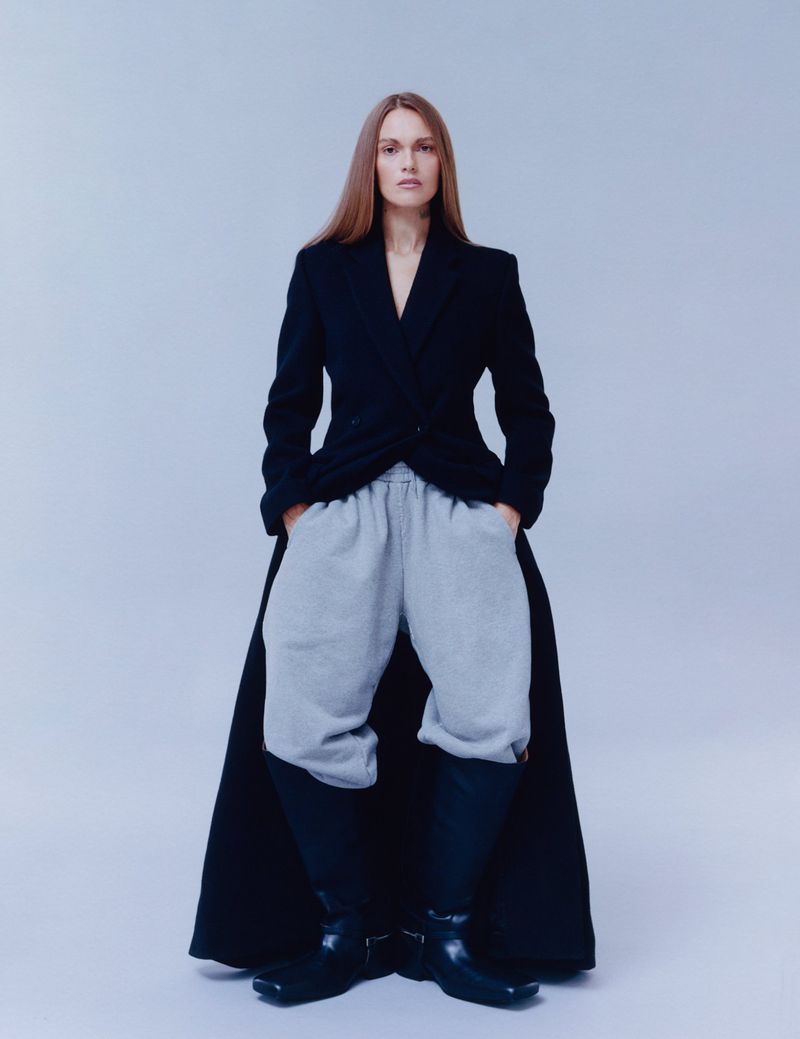 A full-length studio shot of a slender model with long brown hair, wearing a black blazer, grey sweatpants, and black leather boots.