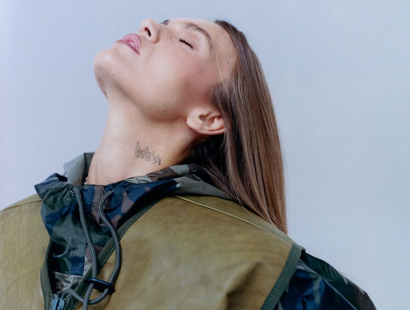 Portrait of an adult female with light skin and blonde hair, tilting her head back to reveal a neck tattoo, wearing a camouflage vest.