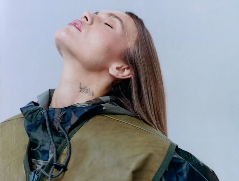 Portrait of an adult female with light skin and blonde hair, tilting her head back to reveal a neck tattoo, wearing a camouflage vest.