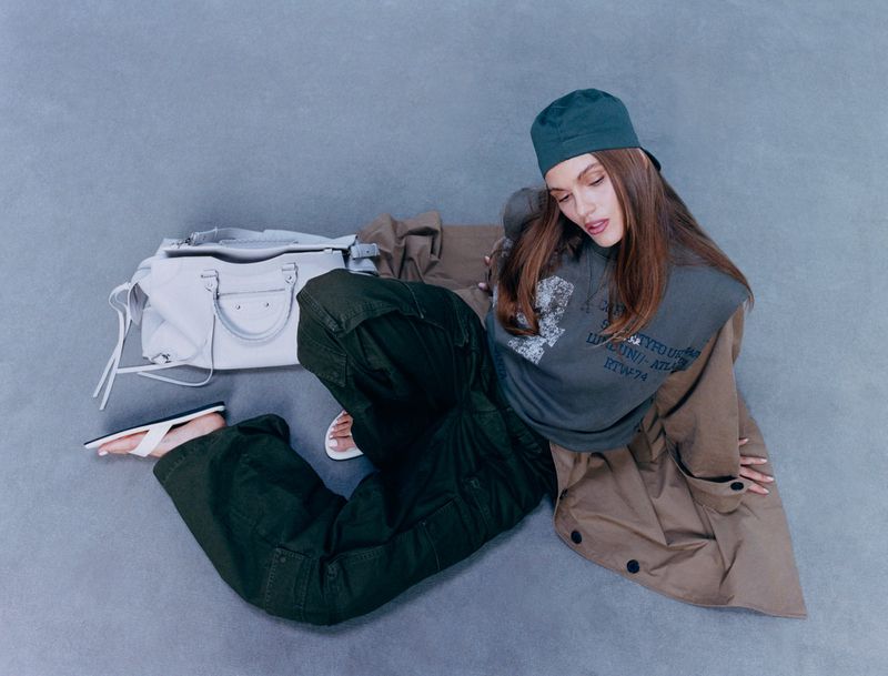 Brunette woman posing with a cap, dark green cargo pants, oversized grey printed t-shirt, tan trench coat and white leather bag.