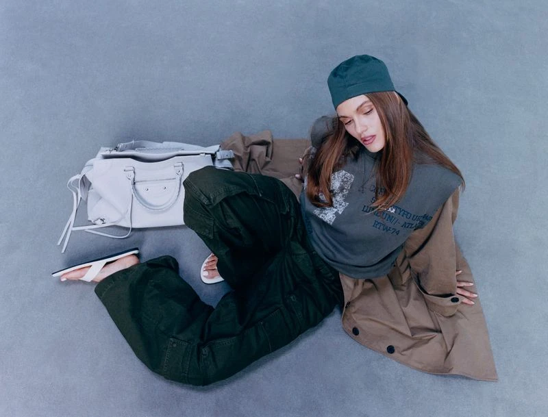 Brunette woman posing with a cap, dark green cargo pants, oversized grey printed t-shirt, tan trench coat and white leather bag.