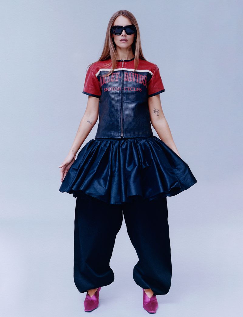 A fashionable woman poses indoors wearing a red, black, and white motorcycle-themed top, voluminous black skirt, black pants, sunglasses, and pink heels.