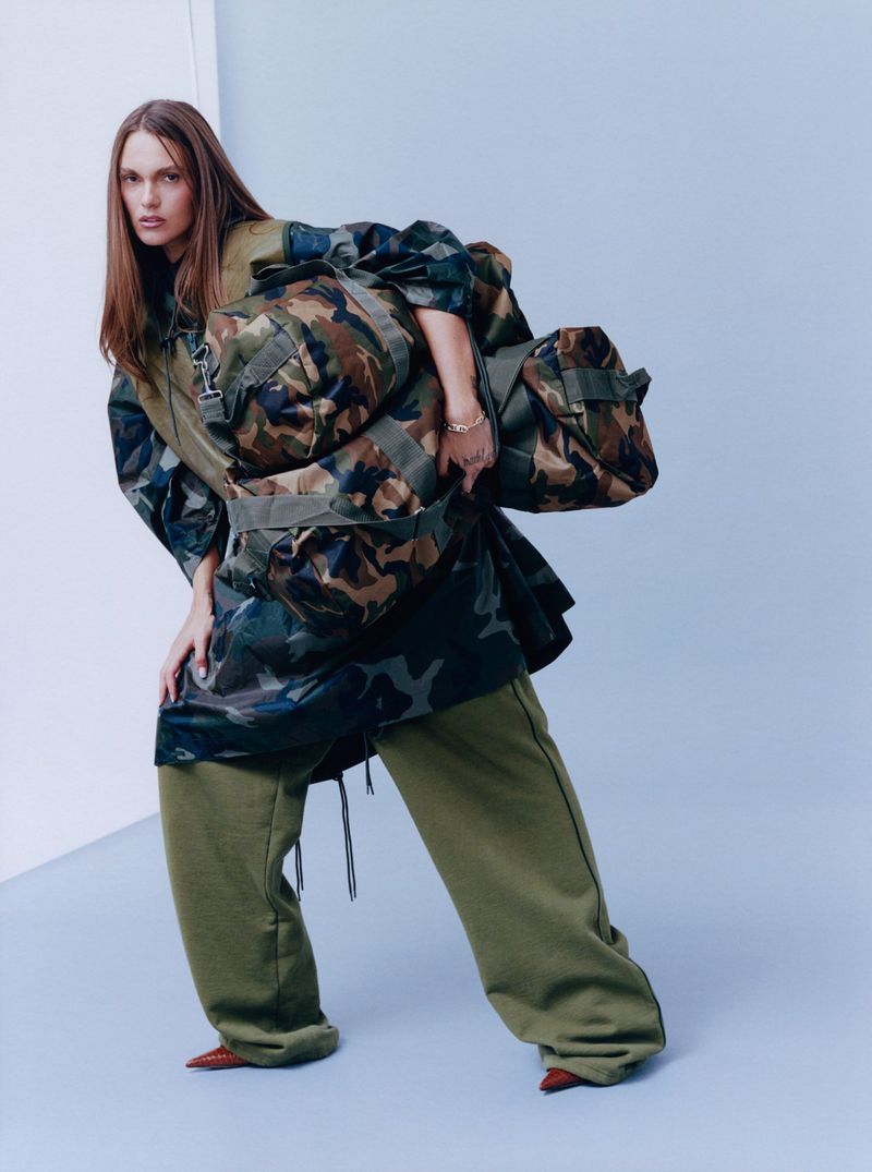 Fashion shot of a model wearing a camouflage jacket and olive sweatpants with a duffel bag in a studio with a gradient light blue background.