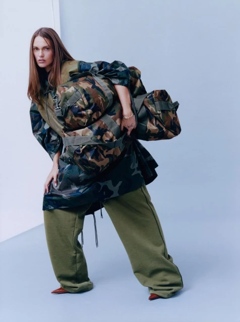 Fashion shot of a model wearing a camouflage jacket and olive sweatpants with a duffel bag in a studio with a gradient light blue background.